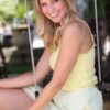abbey-saunders-4 A smiling blonde woman wearing a yellow top sits on a garden swing in bright sunlight.