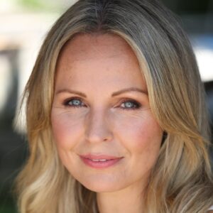 A blonde woman with blue eyes smiling warmly at the camera in a professional commercial headshot portrait.