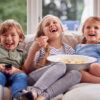 Three Children Sitting On Sofa At Home Laughing And Watching TV With Popcorn