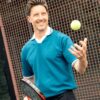 A smiling man holding a padel racket and tossing a tennis ball in the air outdoors.