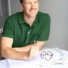 A smiling man in a green polo shirt sits at a desk with paperwork and reading glasses.