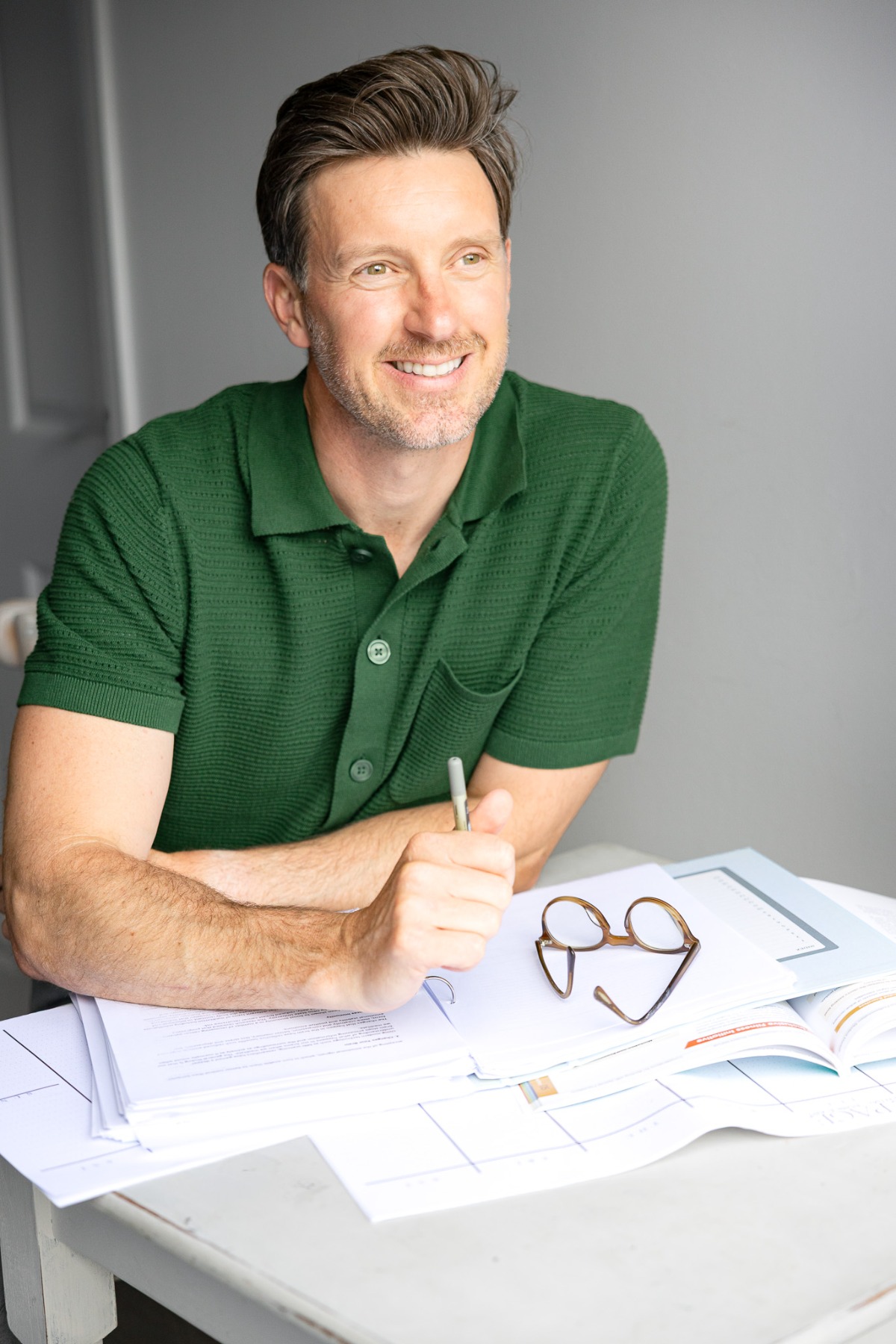 ben-davis-2 A smiling man in a green polo shirt sits at a desk with paperwork and reading glasses.