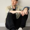 A relatable man in a denim shirt and black trousers sits against a neutral wall looking away.
