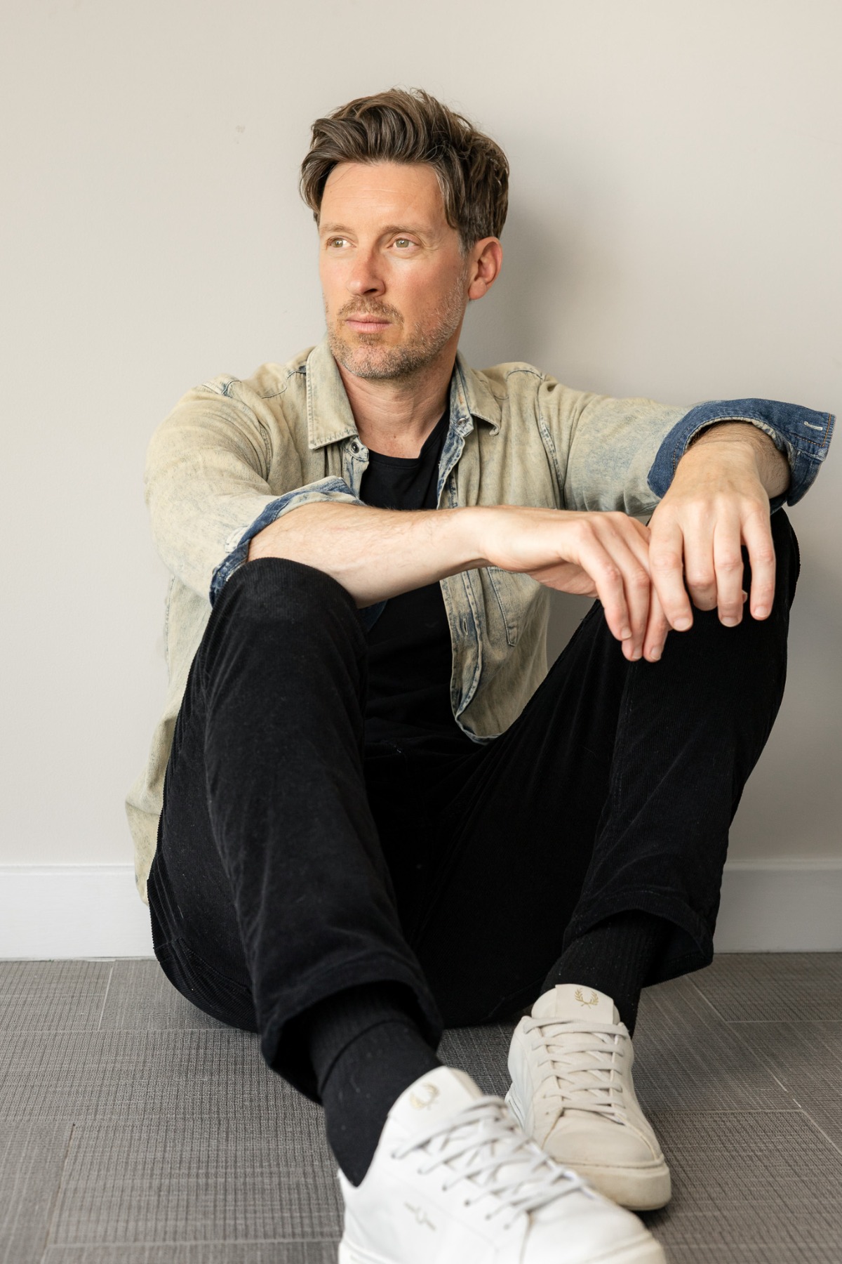 ben-davis-4 A relatable man in a denim shirt and black trousers sits against a neutral wall looking away.