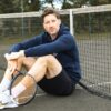 A man in a navy hoodie sits on a tennis court holding a racket looking at camera.