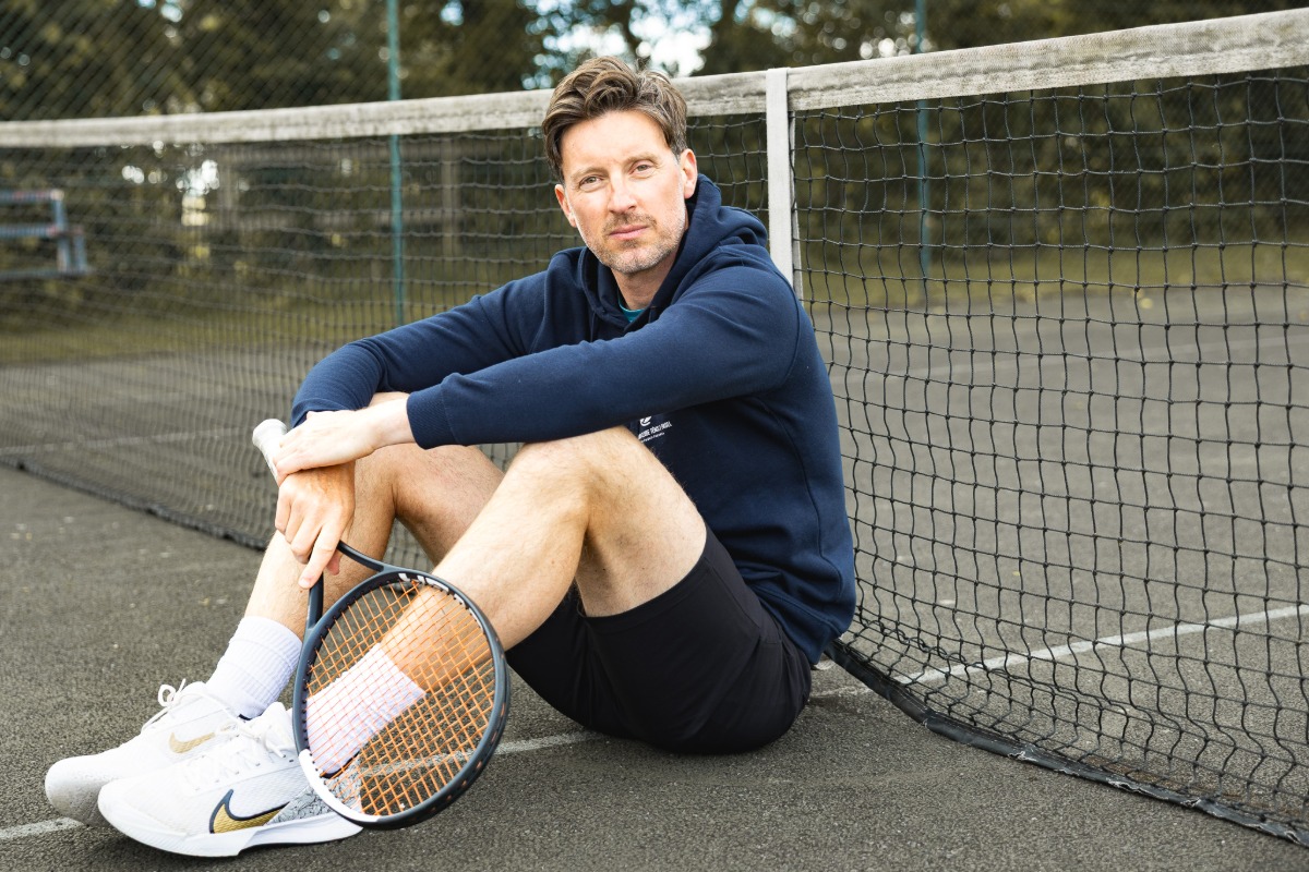 ben-davis-9 A man in a navy hoodie sits on a tennis court holding a racket looking at camera.