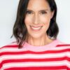 A friendly brunette woman with a bright smile wearing a red and pink striped jumper against white.