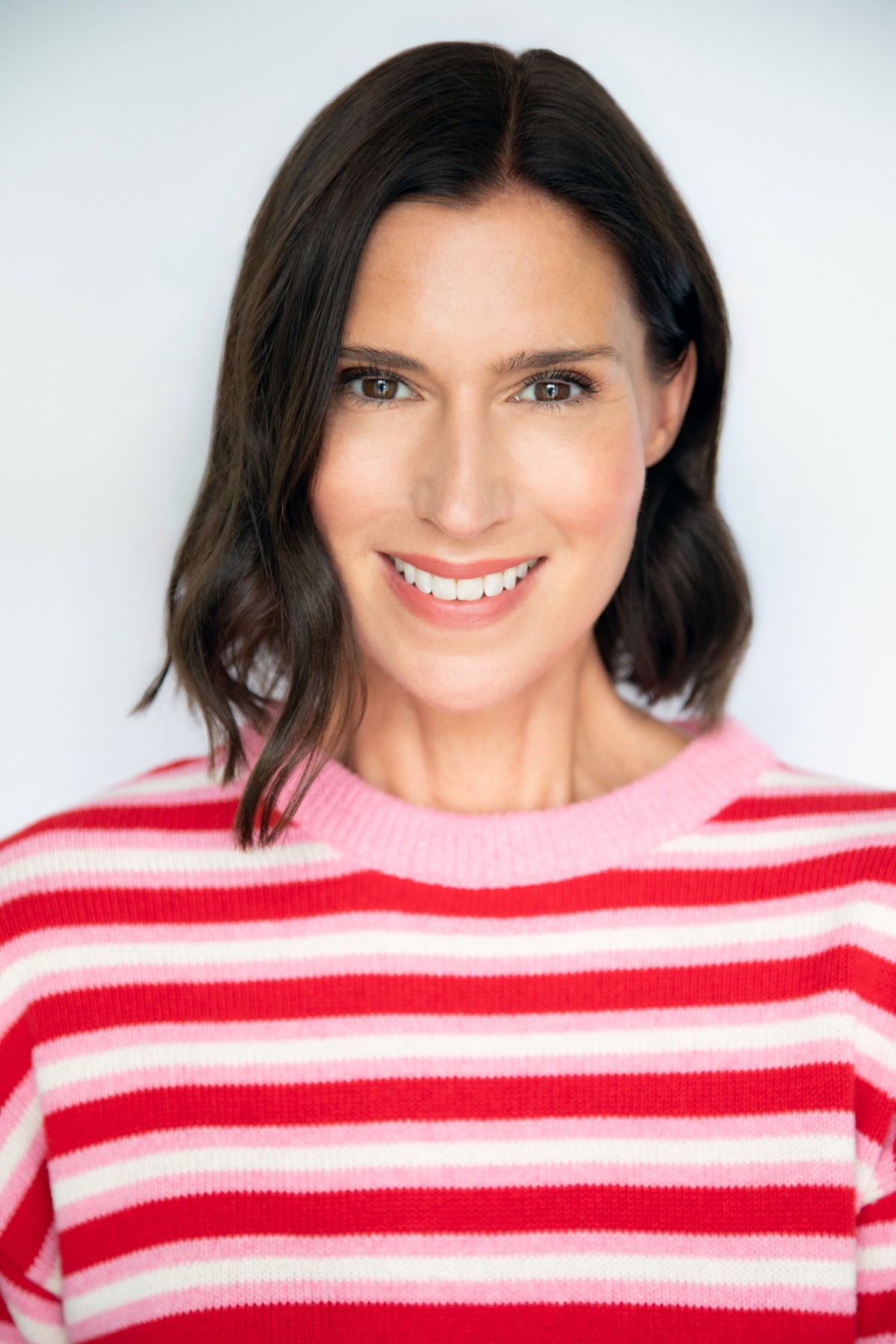 A friendly brunette woman with a bright smile wearing a red and pink striped jumper against white.