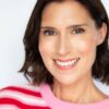 A beaming woman with dark bobbed hair wearing a bright pink and red striped sweater, studio background.