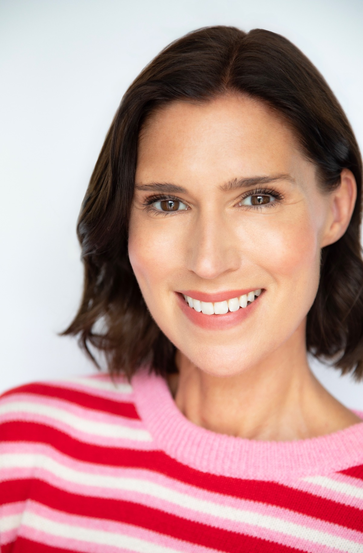 A beaming woman with dark bobbed hair wearing a bright pink and red striped sweater, studio background.