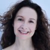 deborah-aharoni-2 A smiling woman with long curly brown hair and a white top posing against a plain background.