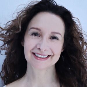 A smiling woman with long curly brown hair and a white top posing against a plain background.