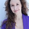 deborah-aharoni-5 A mature woman with curly dark hair wearing a vibrant purple blazer against a plain white background.