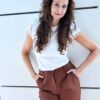 deborah-aharoni-8 A smiling woman with curly brown hair wearing a white top stands against a clean outdoor wall.