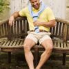 A smiling man sits on a wooden garden bench wearing a yellow polo shirt and light-blue sweater.