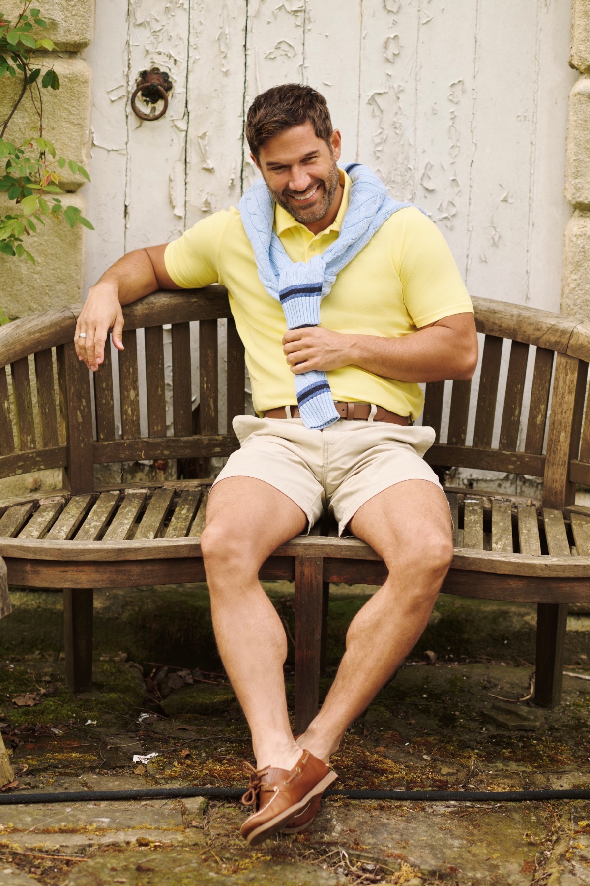 A smiling man sits on a wooden garden bench wearing a yellow polo shirt and light-blue sweater.