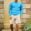 A man wearing a bright blue cable knit sweater and beige shorts walks on a stone garden path.