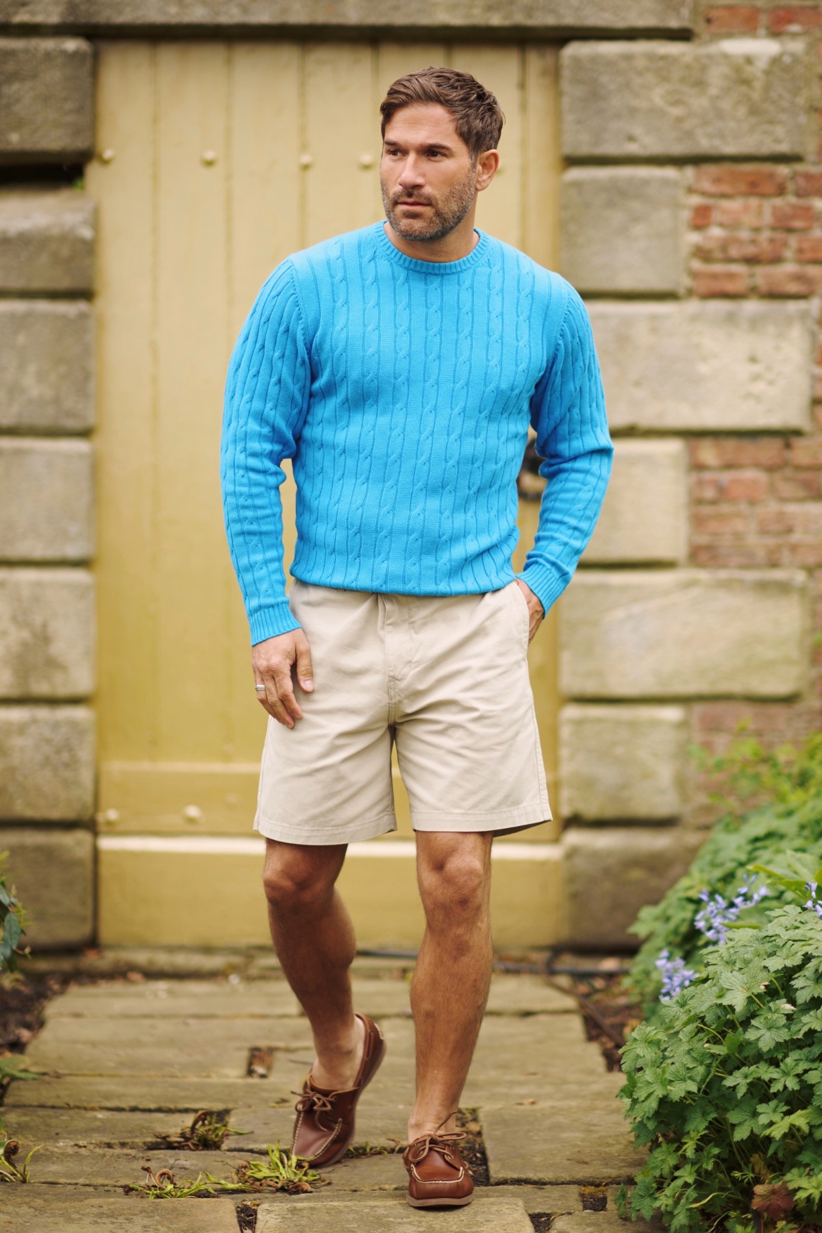 A man wearing a bright blue cable knit sweater and beige shorts walks on a stone garden path.