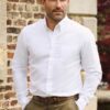 A well-groomed man in a crisp white shirt stands confidently against a rustic brick wall background.