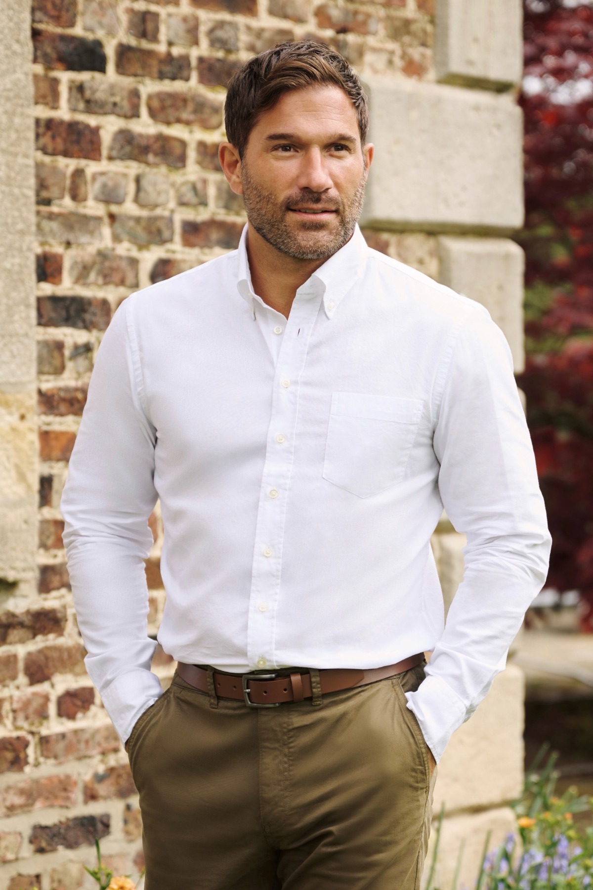 A well-groomed man in a crisp white shirt stands confidently against a rustic brick wall background.