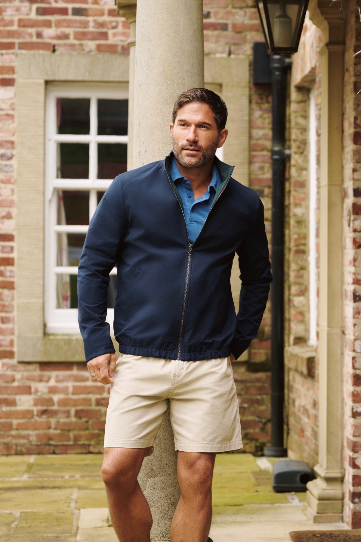 A mature man in a navy jacket and beige shorts standing outdoors in a garden setting.