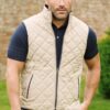 A man wearing a quilted beige gilet and dark polo shirt stands in a lush garden setting.