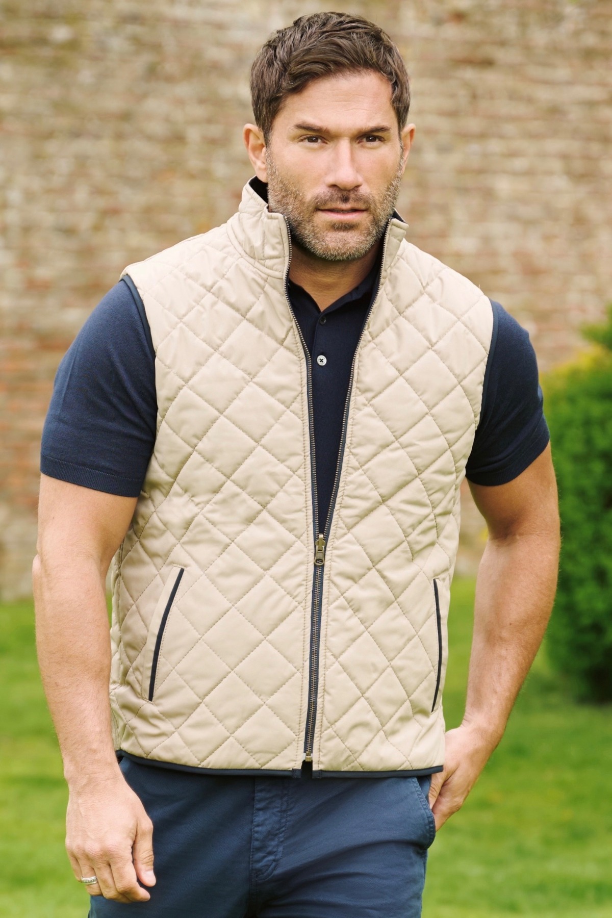 A man wearing a quilted beige gilet and dark polo shirt stands in a lush garden setting.