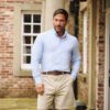 A handsome man in a light blue shirt and beige shorts poses confidently by a brick house pillar.
