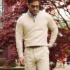 A stylish man wearing a beige quarter-zip sweater and shorts, standing before vibrant red Japanese maple leaves.