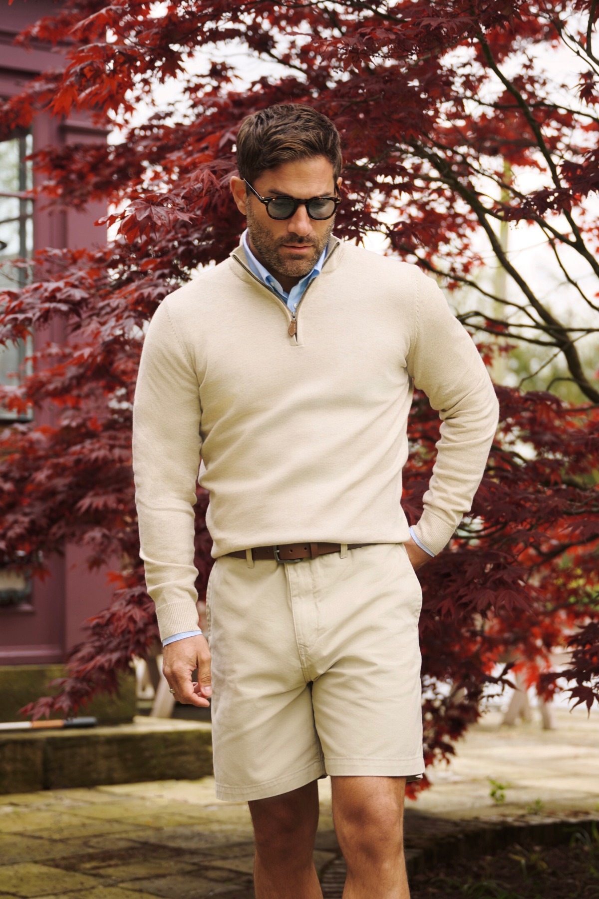 A stylish man wearing a beige quarter-zip sweater and shorts, standing before vibrant red Japanese maple leaves.