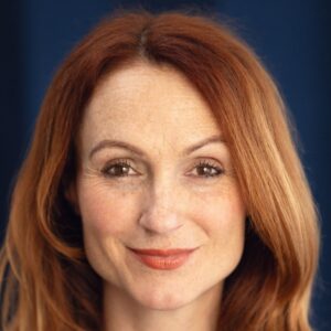 A warm, smiling ginger-haired woman with freckles poses for a clean and professional studio headshot portrait.
