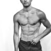 A fit shirtless male model with short hair stands against a plain white studio background.