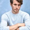 A man with curly brown hair wearing a light blue sweater poses against a blue textured studio backdrop.