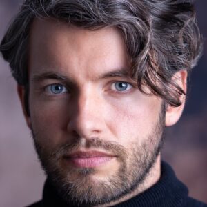 A man with blue eyes and salt and pepper hair looking directly into the camera lens indoors.