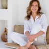 A smiling mature woman in white linen casual clothing sits cross-legged against a bright, warm textured wall.
