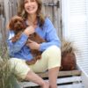 A smiling woman in a blue sweater sits outdoors on wooden steps holding her small brown dog.