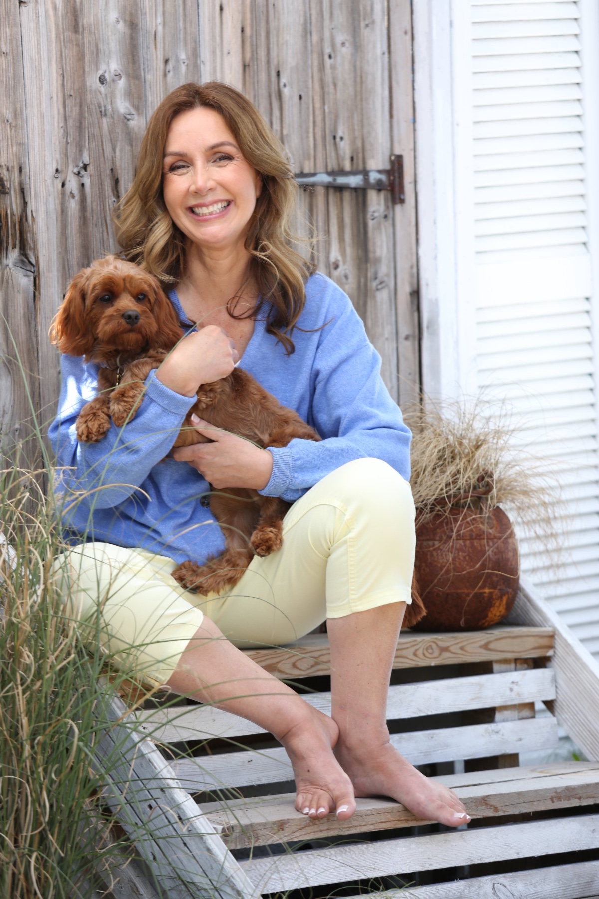 A smiling woman in a blue sweater sits outdoors on wooden steps holding her small brown dog.