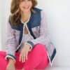 A smiling mature woman sits against a white wall wearing a stylish tweed jacket and pink trousers.