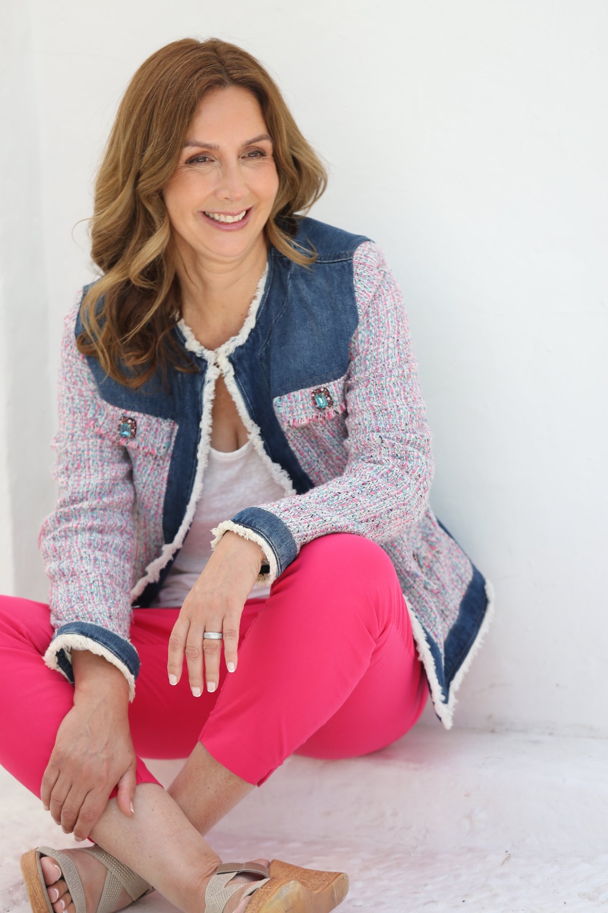 A smiling mature woman sits against a white wall wearing a stylish tweed jacket and pink trousers.
