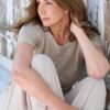 A mature woman with long brunette hair poses thoughtfully against a rustic weathered wooden wall environment.