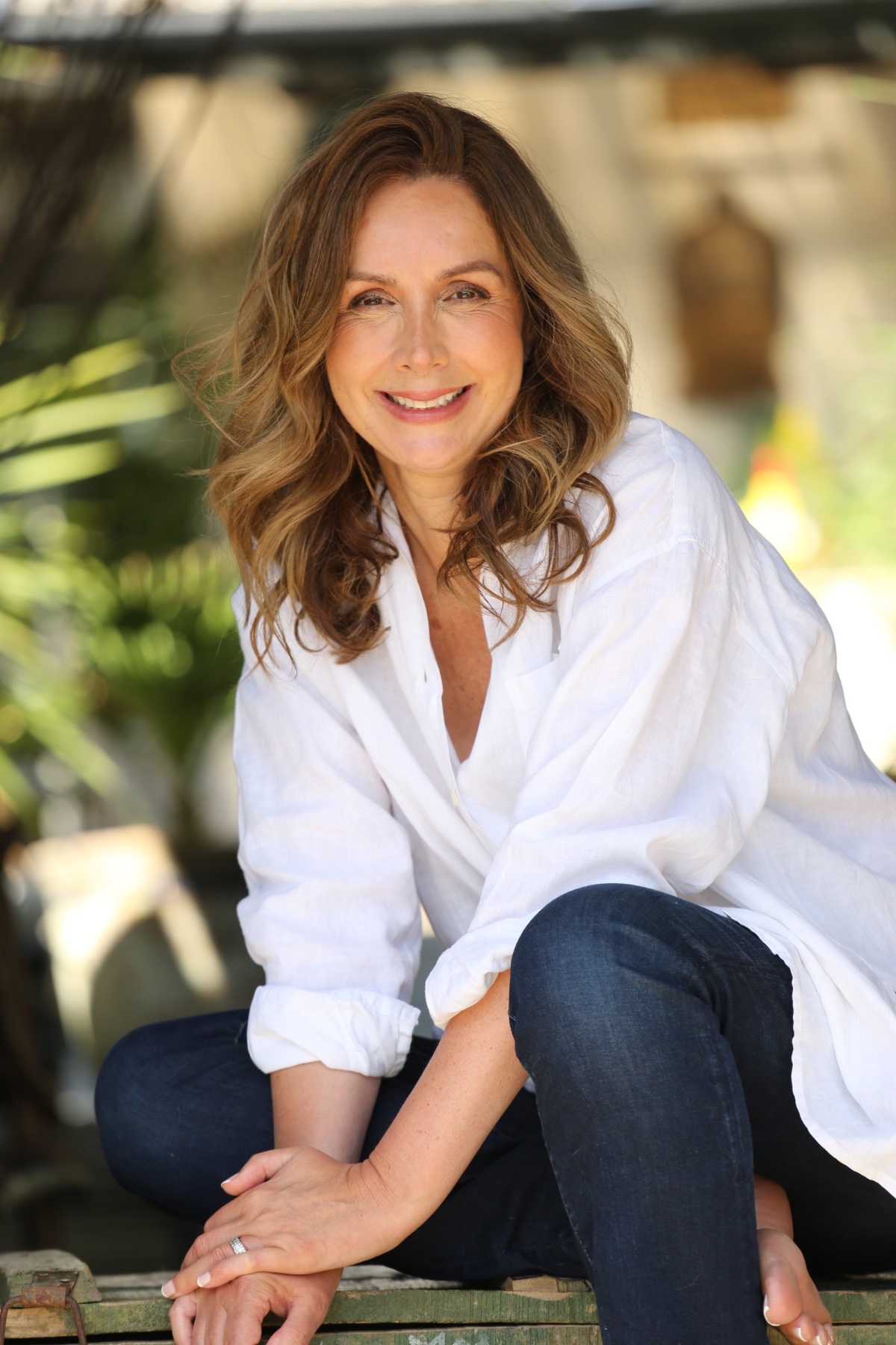 A radiant woman with shoulder-length wavy hair smiling naturally towards the camera in a bright outdoor setting.