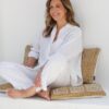 A smiling woman with wavy hair in white linen clothing sits comfortably against a textured white wall.