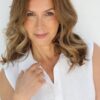 A warm portrait of a mature woman with wavy brown hair wearing a simple white sleeveless shirt.