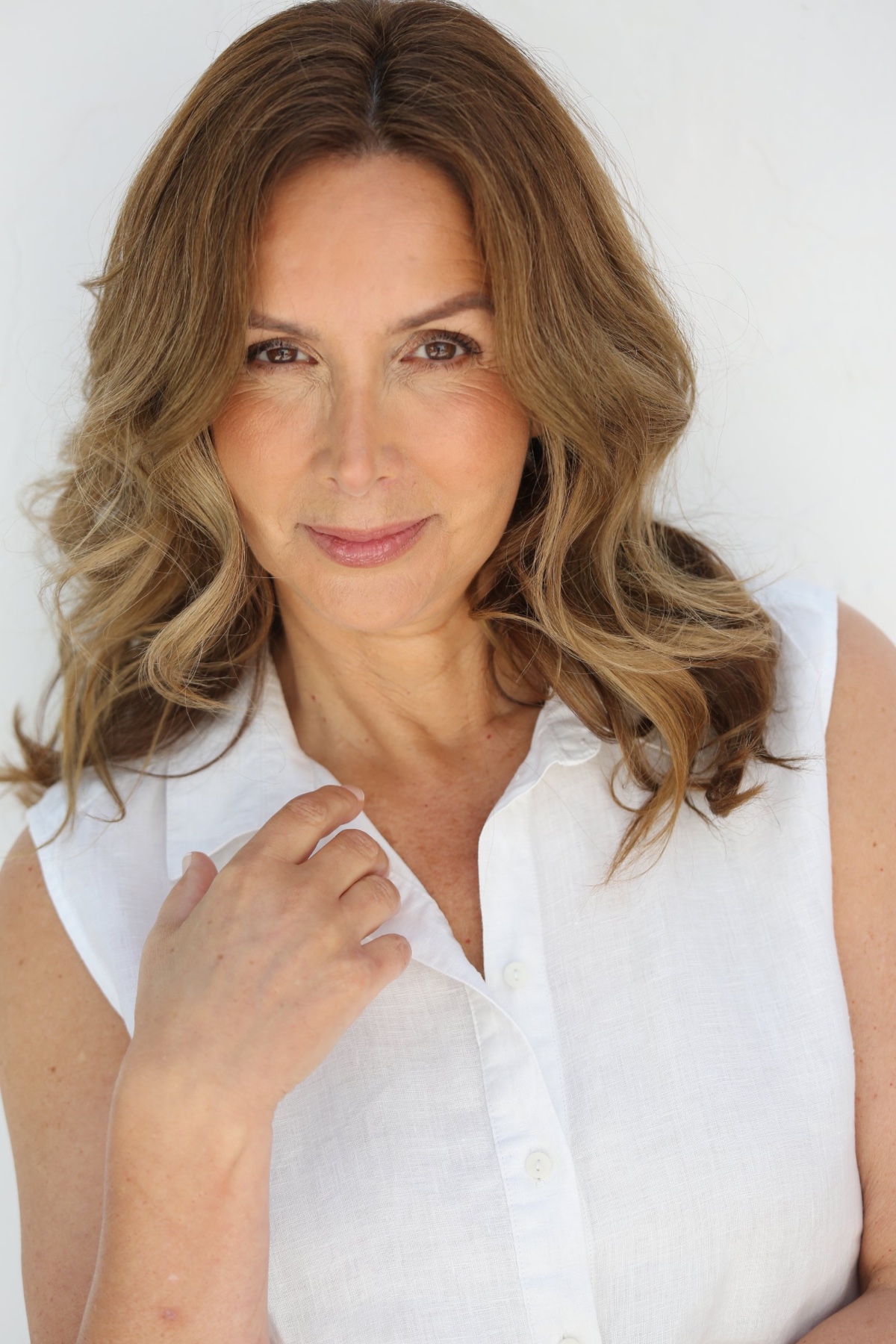 A warm portrait of a mature woman with wavy brown hair wearing a simple white sleeveless shirt.
