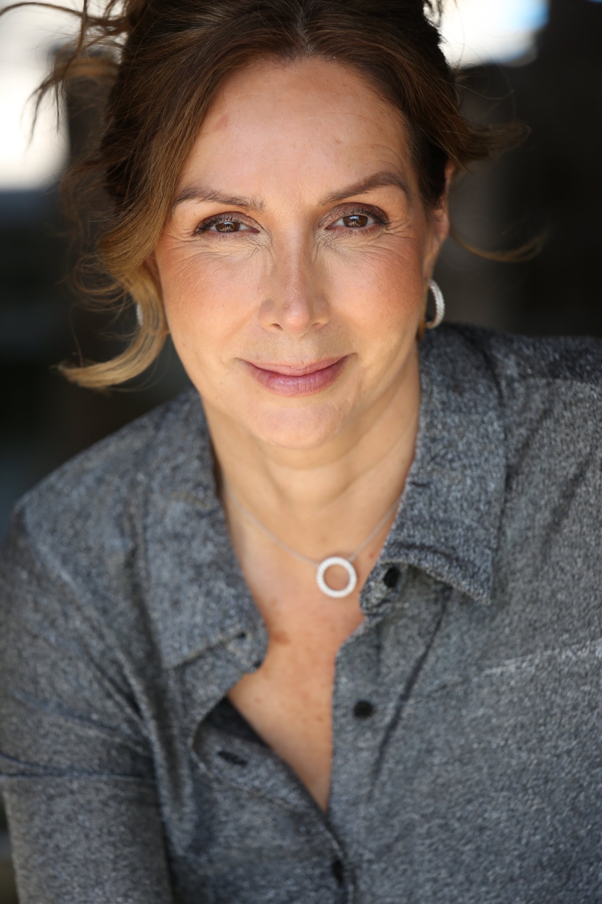 Mature woman with a warm, friendly smile looks directly into the camera in a soft natural light portrait.