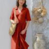 carol-ann-dunbar-8 A smiling mature woman in a rust-coloured dress stands against a white wall with Mediterranean decor accents.