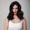 A dark-haired woman with wavy hair poses for a professional studio headshot against a neutral grey background.