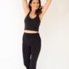 A smiling mature woman in black activewear stretches her arms overhead against a clean white studio background.