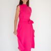 A smiling brunette woman wearing a bright pink dress stands against a clean white studio background.