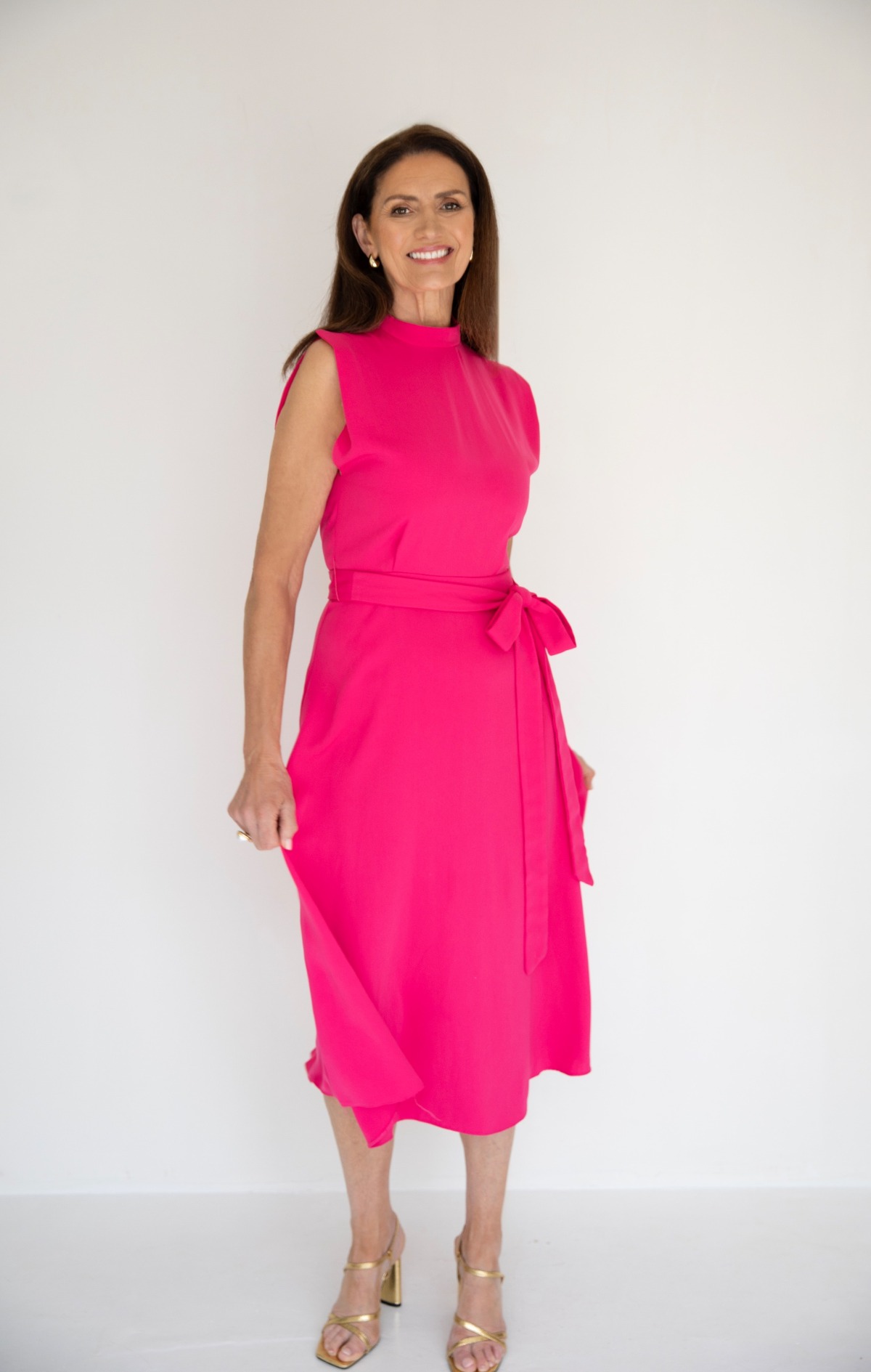 A smiling brunette woman wearing a bright pink dress stands against a clean white studio background.